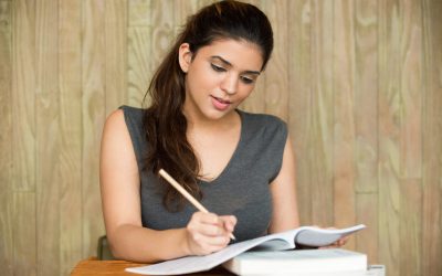 Portrait of young Caucasian female student sitting at desk, writing in copybook and smiling in classroom